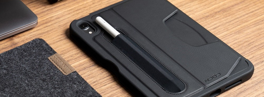 A purple iPad case with grooves and an Apple Pencil is displayed on a flat surface surrounded by accessories