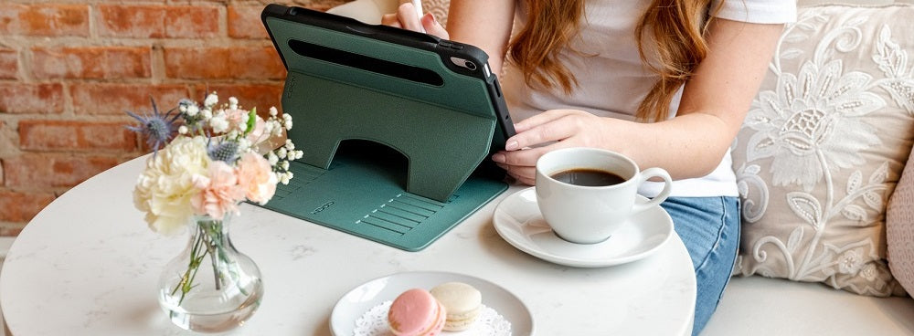 A person with long red hair is using a tablet on a marble table with a cup of coffee, macarons, and a vase of flowers. Brick wall and cushions are in the background.