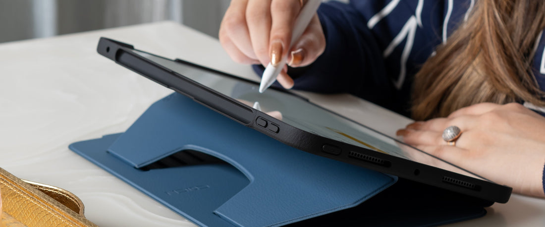 An overhead, close-up shot shows a person's hands using a white stylus on a tablet that's propped up by a blue Zugu iPad case on a white table.