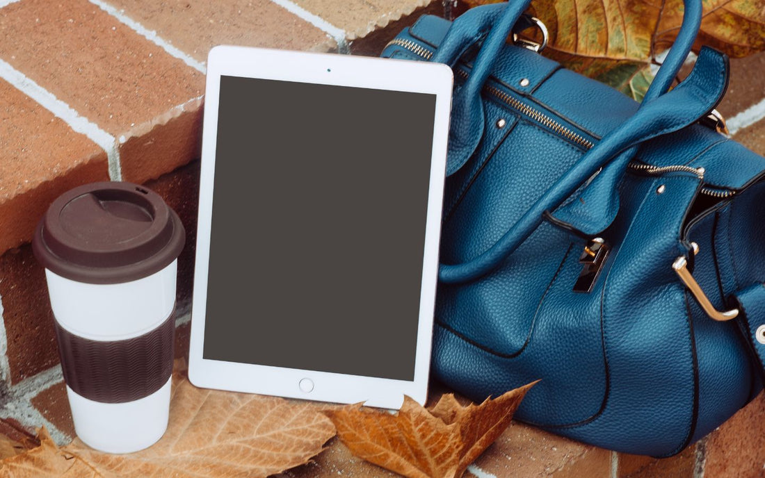 An iPad rests on a blue handbag next to a takeout coffee cup