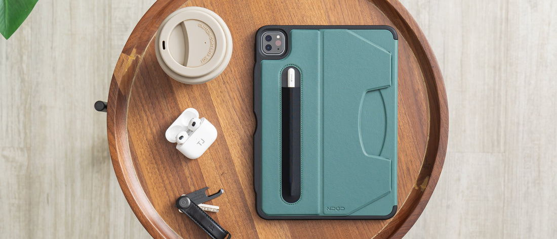A round wooden table, with an iPad in a ZUGU case in green, a USB stick, Apple AirPods and a cup of coffee resting on it. 