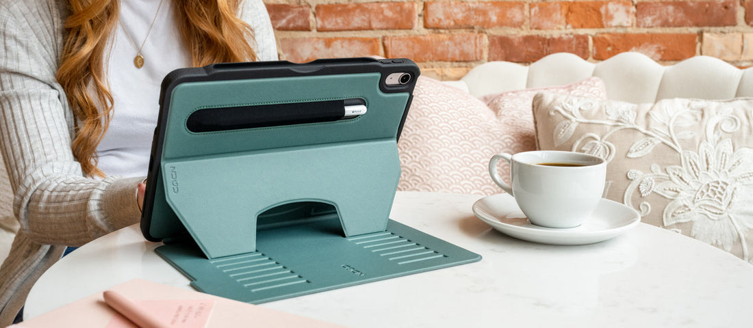 Back view of an iPad in a teal ZUGU case with adjustable stand open and Apple Pencil stored in the groove, on a café table beside a cup of tea 