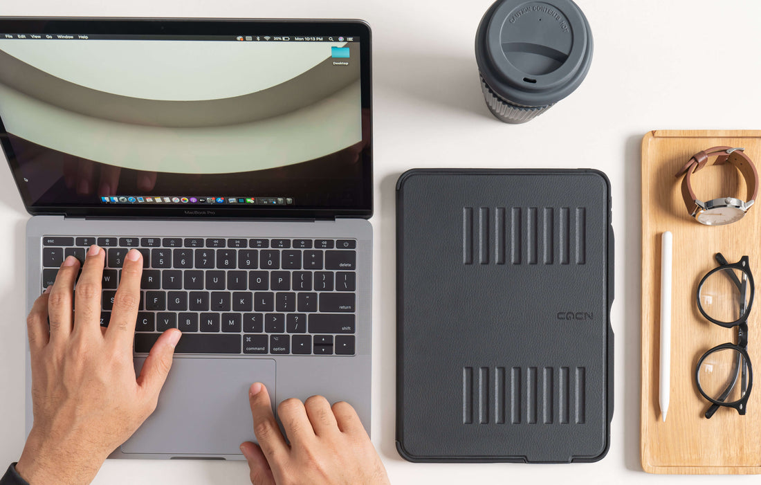 A man using a laptop, with an iPad in a black ZUGU case on the table on the right. A cup of coffee, a pair of glasses, and an Apple Pencil are also displayed on the table.