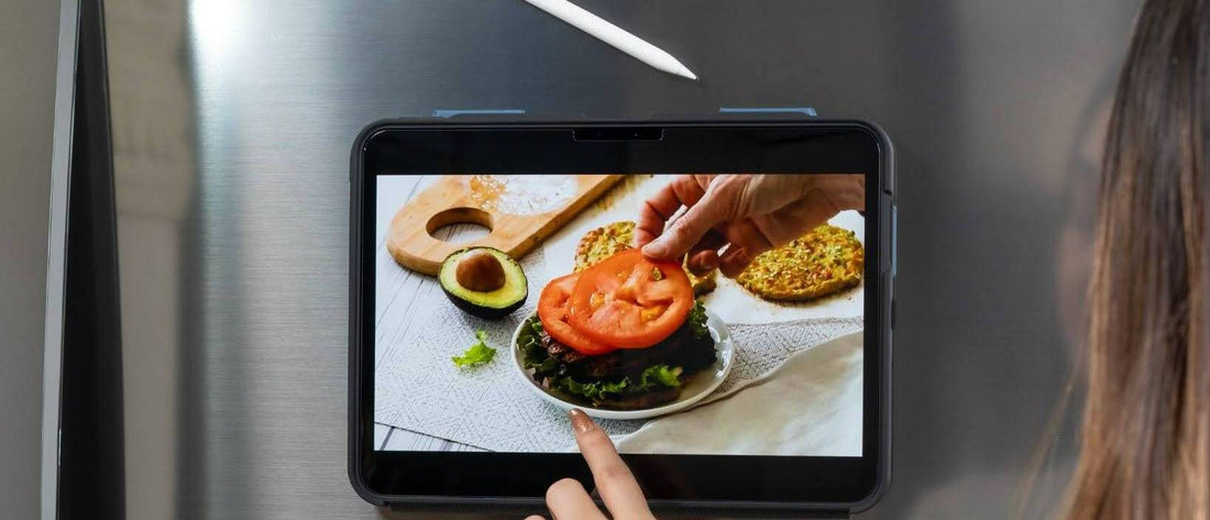 iPad in a ZUGU case magnetically mounted on a stainless steel fridge, with a woman tapping through a recipe photo hands-free while cooking